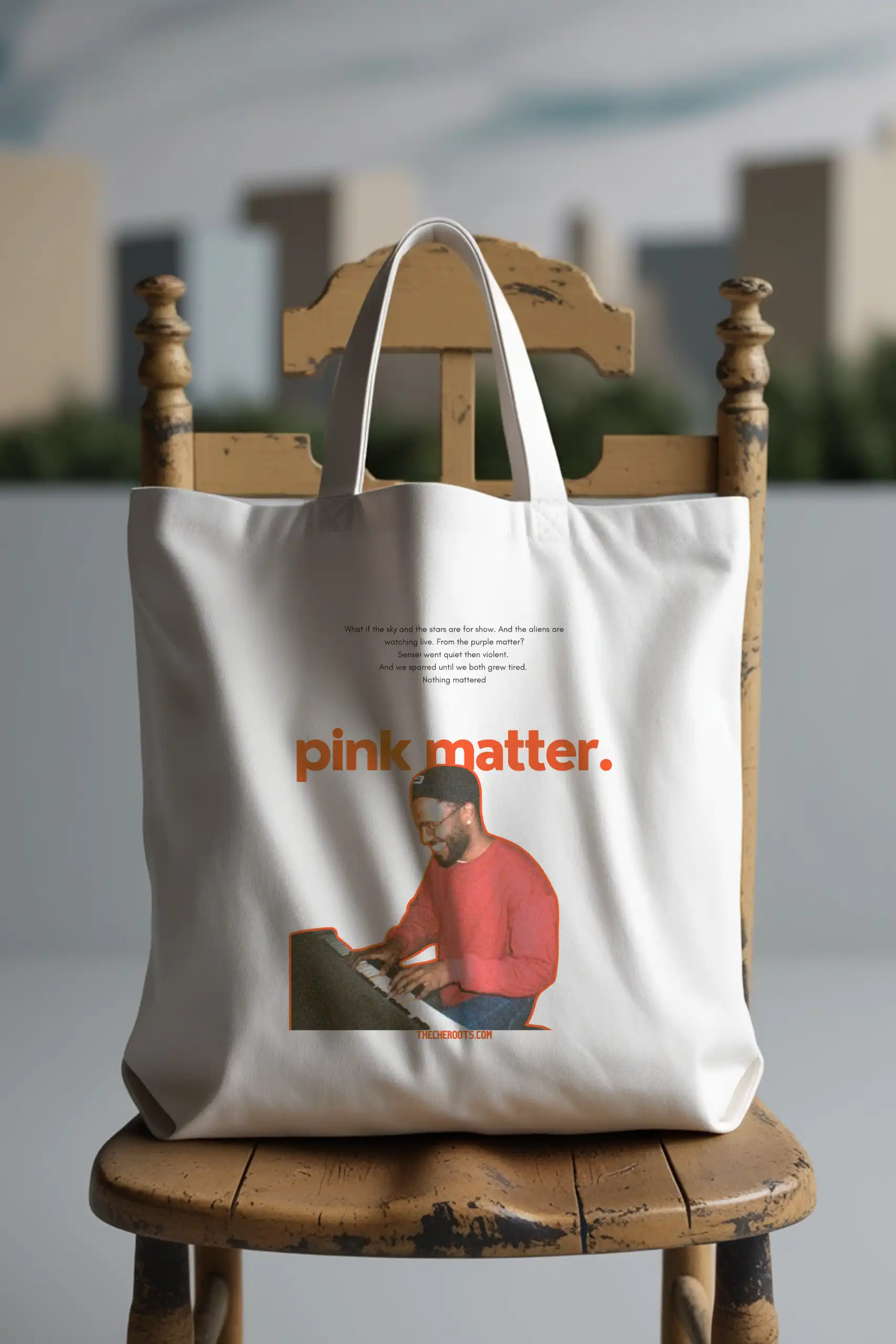 A white tote bag with the text "pink matter." printed on it, along with a black and white image of Frank Ocean playing the piano. The words "THECHEROOTS.COM" are also included. The tote bag is sitting on a wooden chair.