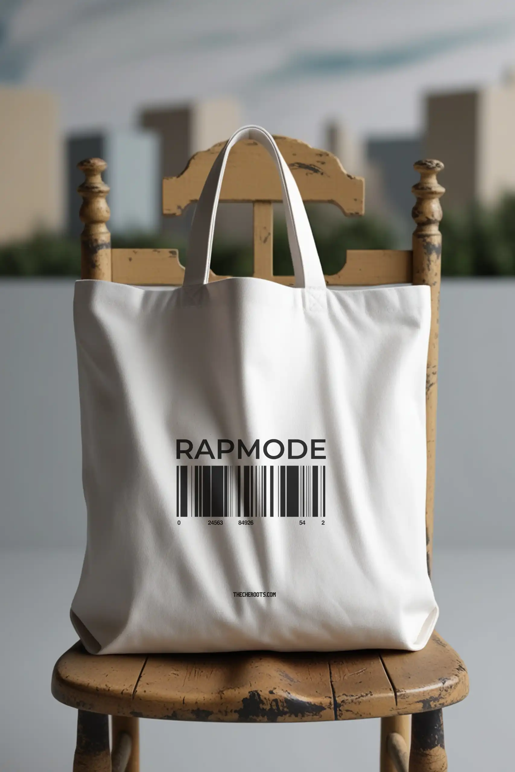 A white tote bag with the text "RAPMODE" printed on it, along with a barcode underneath. The words "THECHEROOTS.COM" are also included. The tote bag is sitting on a wooden chair.