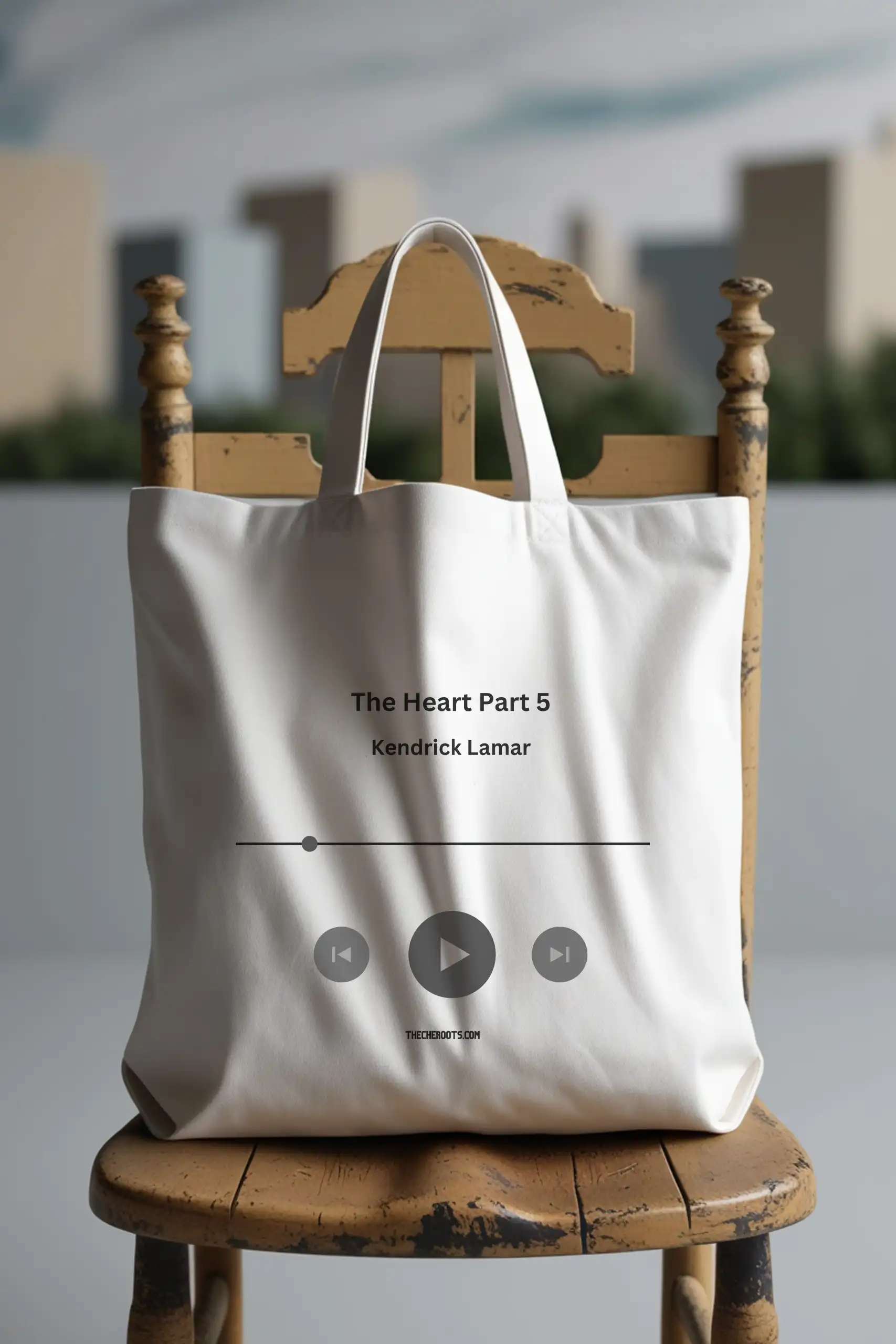 A white tote bag with the text "The Heart Part 5" and "Kendrick Lamar" printed on it, along with a music player interface with play, pause, and skip buttons. The words "THECHEROOTS.COM" are also included. The tote bag is sitting on a wooden chair.