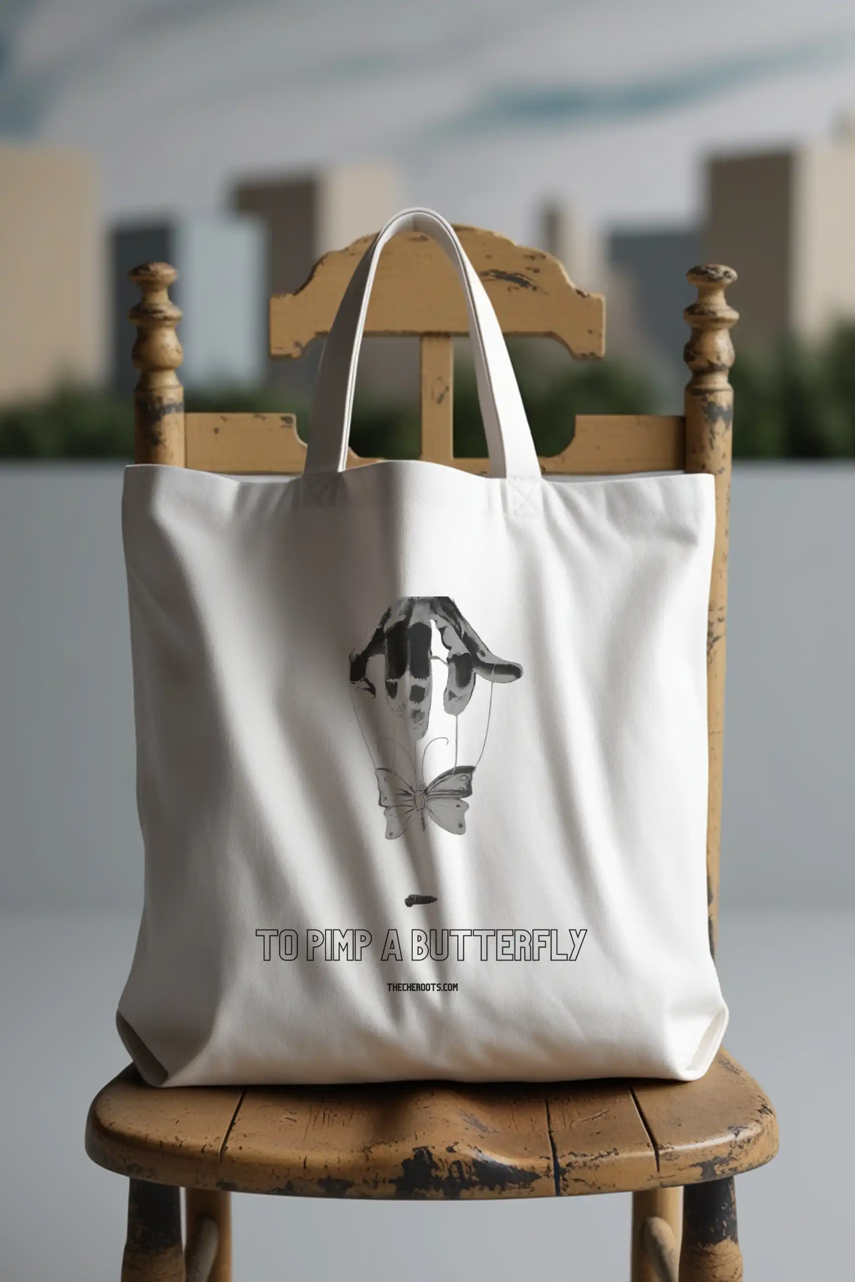 A white tote bag with the text "TO PIMP A BUTTERFLY" printed on it, along with a graphic of a hand holding a butterfly with a ribbon around it. The words "THECHEROOTS.COM" are also included. The tote bag is sitting on a wooden chair.