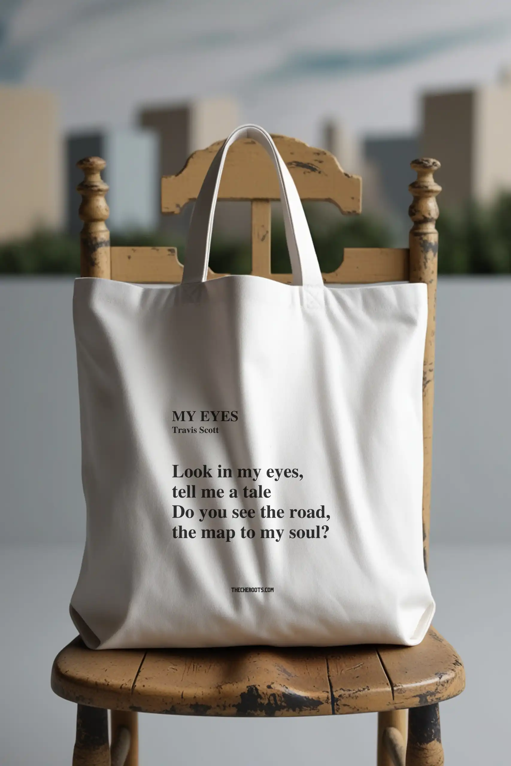 A white tote bag with the text "MY EYES" and a quote by Travis Scott printed on it. The quote reads: "Look in my eyes, tell me a tale. Do you see the road, the map to my soul?" The words "THECHEROOTS.COM" are included below the quote. The tote bag is sitting on a wooden chair.