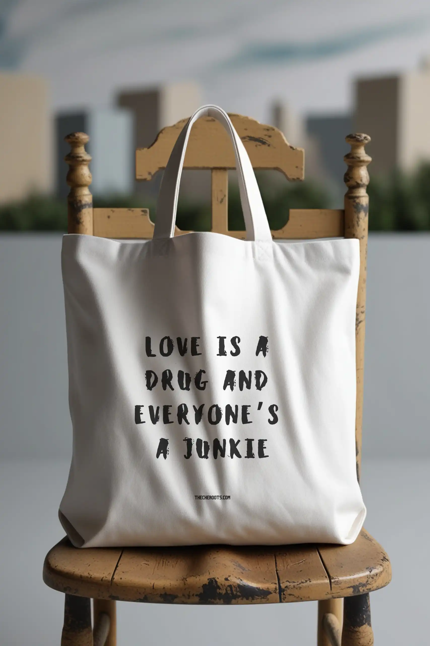 A white tote bag with the text "LOVE IS A DRUG AND EVERYONE'S A JUNKIE" printed in black. The words "THECHEROOTS.COM" are included below the text. The tote bag is sitting on a wooden chair. the background is a city view