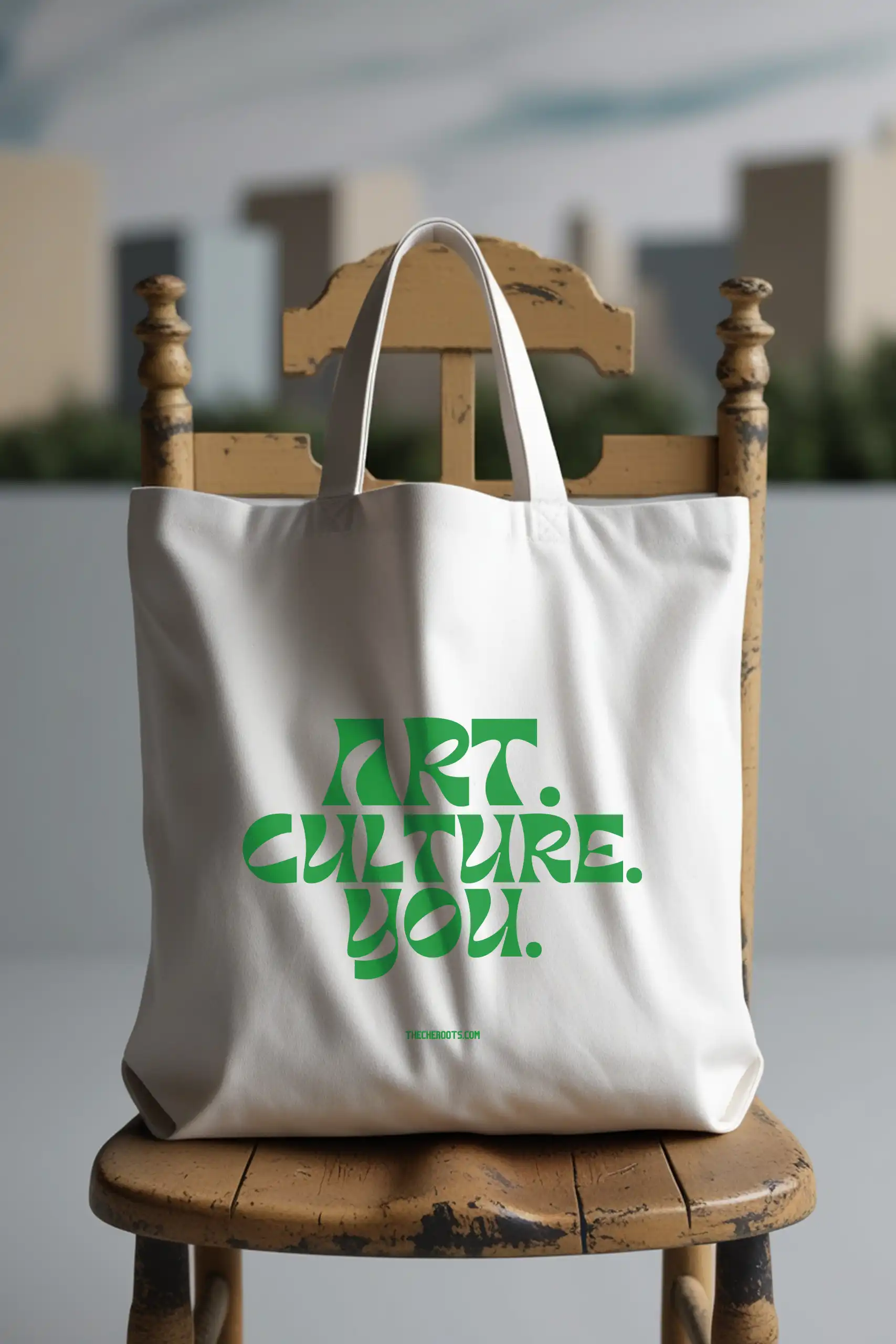 A white tote bag with the text "ART. CULTURE. YOU." printed in green. The words "THECHEROOTS.COM" are included below the text. The tote bag is sitting on a wooden chair.